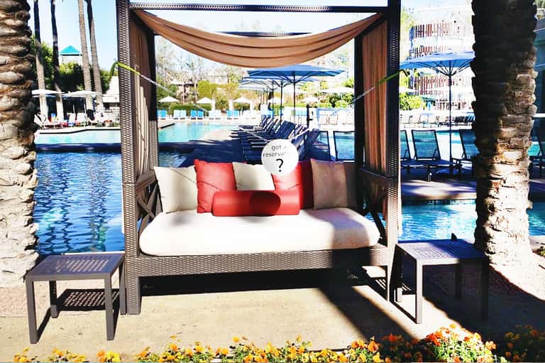 Luxurious poolside cabana bed with brown drapes, red and white pillows, overlooking resort pool and palm trees