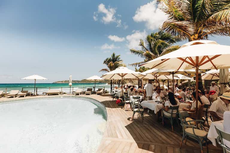 Beachfront restaurant with white umbrellas, palm trees, ocean view, and guests dining al fresco