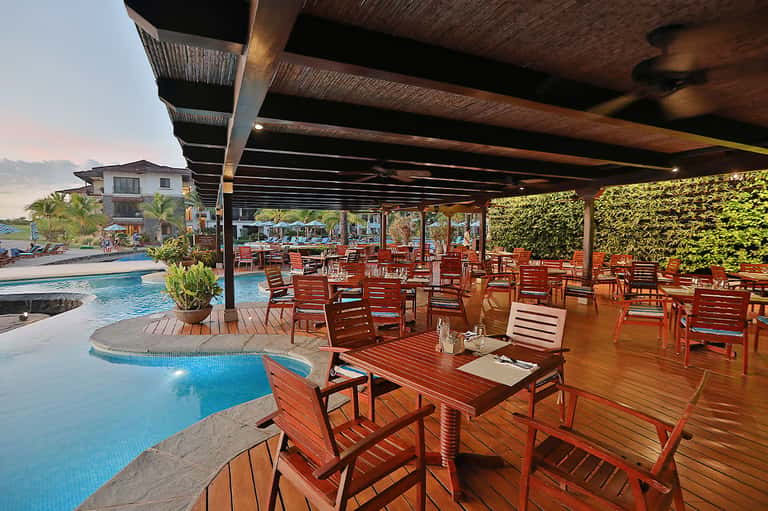 Covered poolside dining area with wooden furniture, tables, pool view, and lush landscaping
