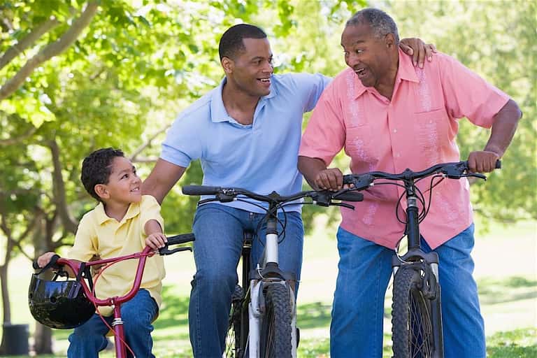 Three generations of men smiling while riding bicycles together outdoors on a tree-lined path
