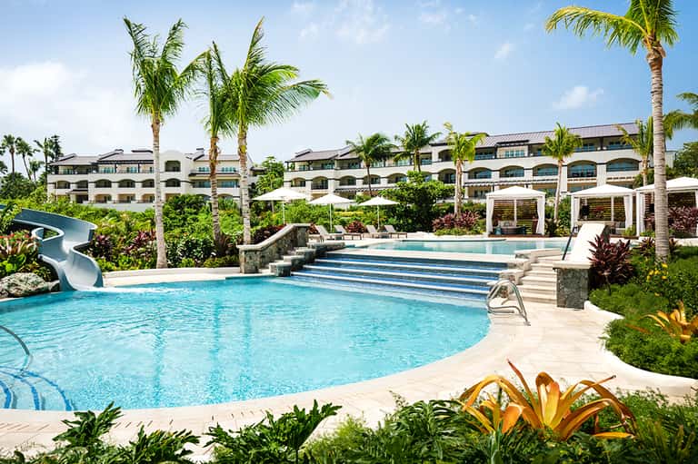 Resort pool complex with water slide, palm trees, lounge areas, and multi-story building
