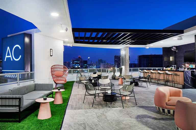 Modern rooftop lounge at dusk with city skyline view, seating areas, bar, and glowing AC sign