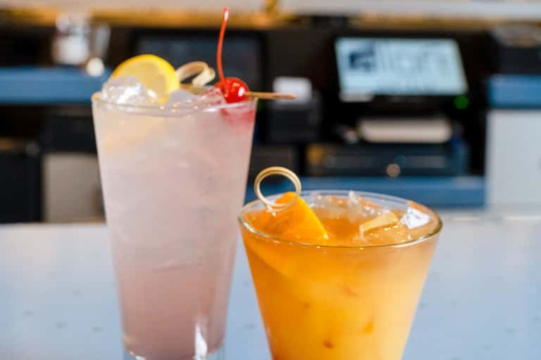 Two tropical cocktails at bar counter with lemon slice, cherry garnish, and blurred background
