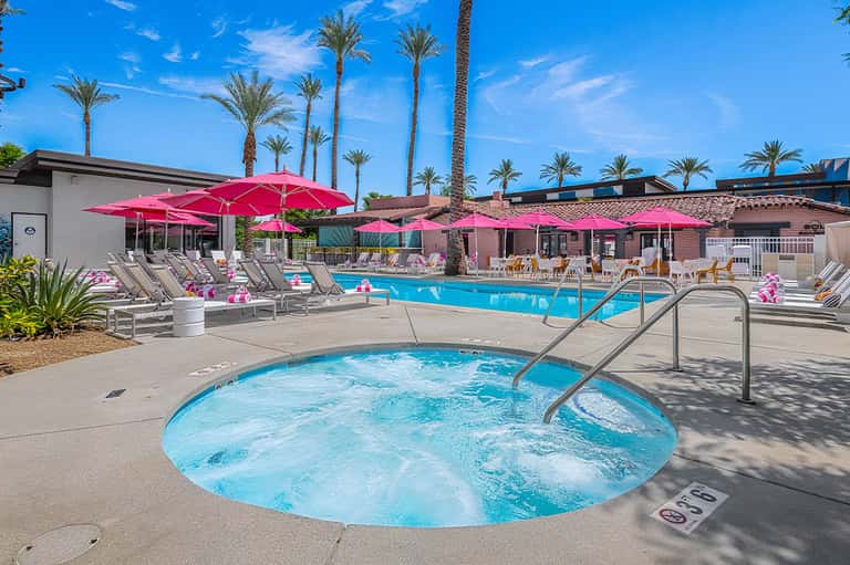 Resort pool complex with hot tub, pink umbrellas, and palm trees under blue sky