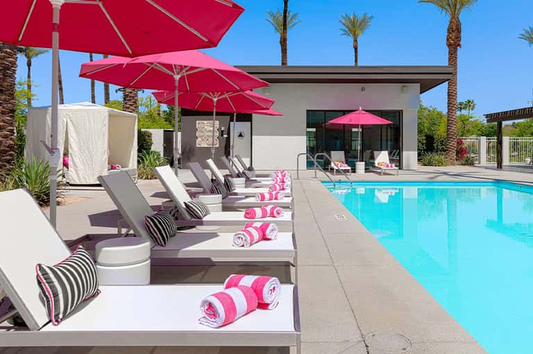 Resort pool area with pink umbrellas, gray lounge chairs, pink towels, and modern spa building under palm trees