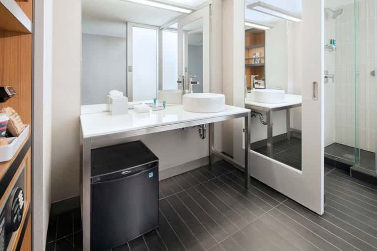 Modern bathroom vanity with dual white sinks, large mirror, black tile floor, and glass shower enclosure.