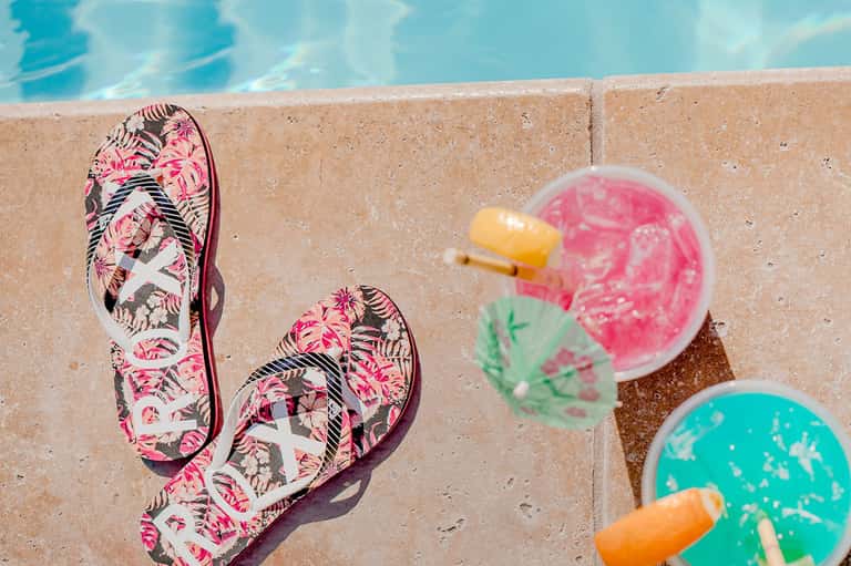 Colorful flip-flops on pool deck next to tropical drinks with umbrellas and pool water