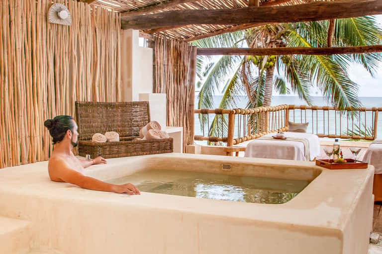 Guest relaxing in outdoor soaking tub on beachfront spa terrace with ocean views and tropical furnishings