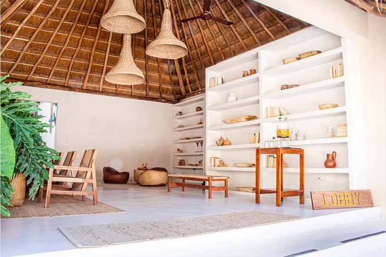 Modern resort lobby with thatched ceiling, white shelving, wooden furniture, and tropical plants