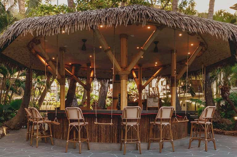 Beachfront tiki bar with thatched roof, wooden railings, woven bar stools, and warm lighting