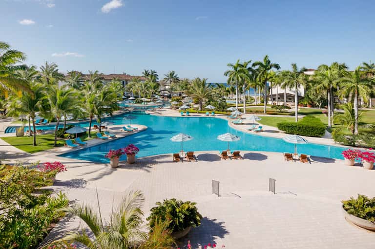 Large resort pool complex with multiple sections, palm trees, and beachfront hotel buildings