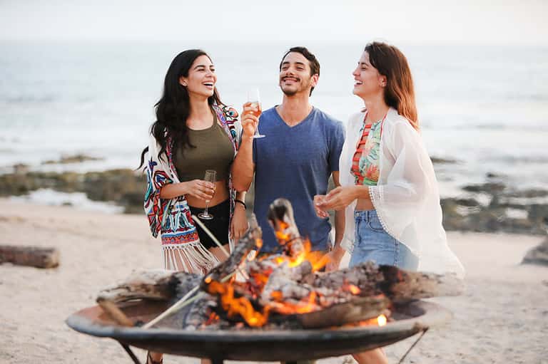 Three friends enjoying champagne by beach bonfire at sunset with ocean and rocky coastline in background