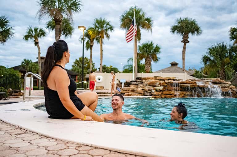 Family enjoying resort pool with waterfall feature, palm trees, and American flag in background