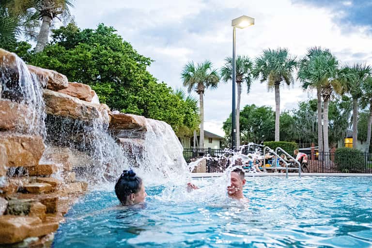 Resort pool with waterfall feature, palm trees, and guests swimming on sunny day