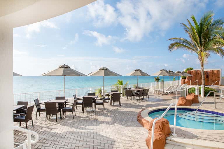 Beachfront terrace with dining tables, umbrellas, ocean view, pool, and palm tree