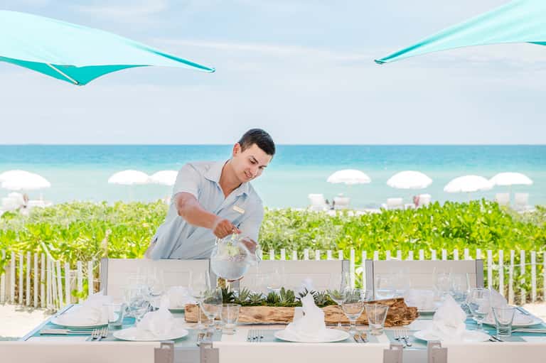 Server pouring water at beachfront dining table with turquoise umbrellas