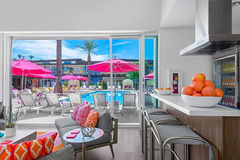 Resort lounge overlooking pool area with pink umbrellas, palm trees, modern furniture, and bright blue sky.