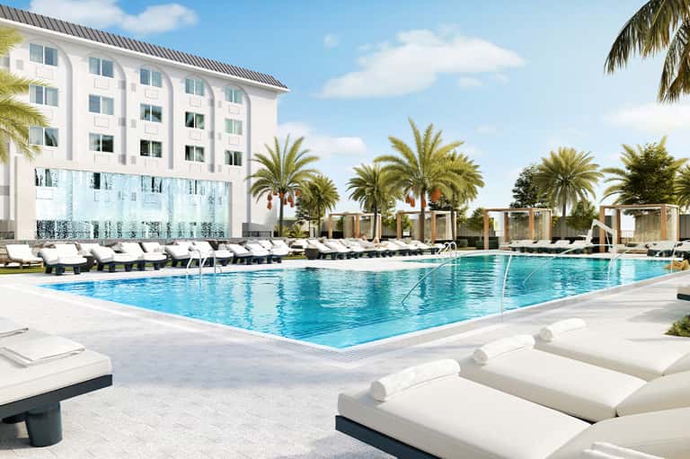 Resort swimming pool with white lounge chairs, palm trees, and modern building with glass wall backdrop