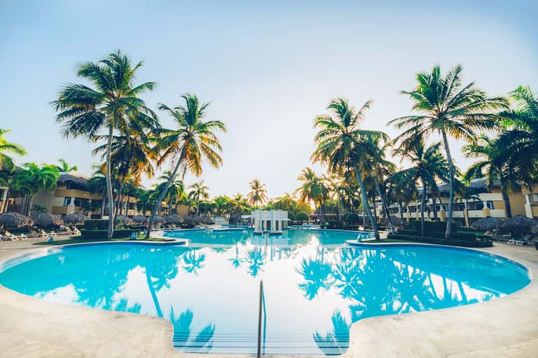 Large turquoise resort pool surrounded by palm trees and beige multi-story hotel buildings