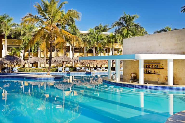 Resort pool with thatched umbrellas, palm trees, lounge chairs, and swim-up bar structure