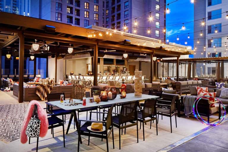 Rooftop lounge area with dining tables, string lights, and buildings at twilight