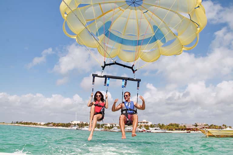 Two people parasailing over turquoise water with yellow parachute and tropical shoreline