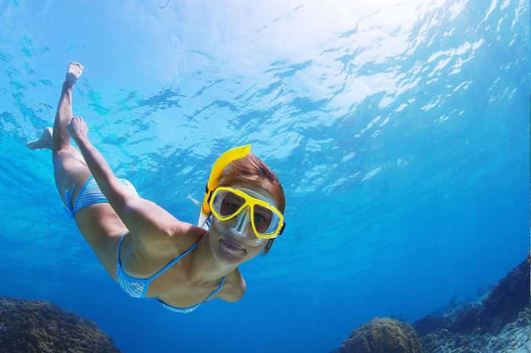 Snorkeler in yellow goggles underwater with coral reef, school of fish and sunlight streaming above