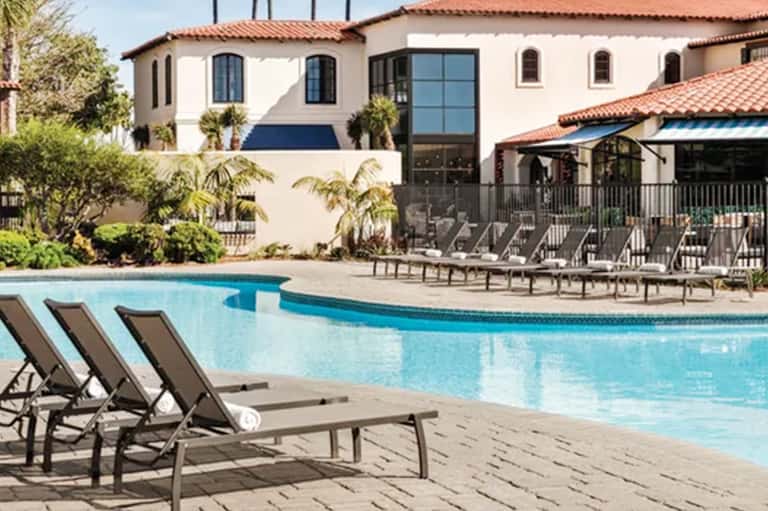 Spanish-style resort pool with lounge chairs, white building, palm trees, and covered patio area