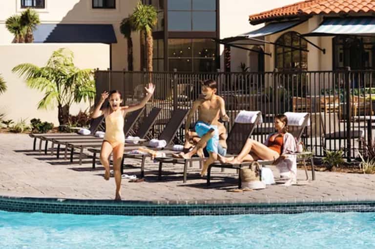 Family playing by resort pool with children, lounge chairs, and Spanish-style roof