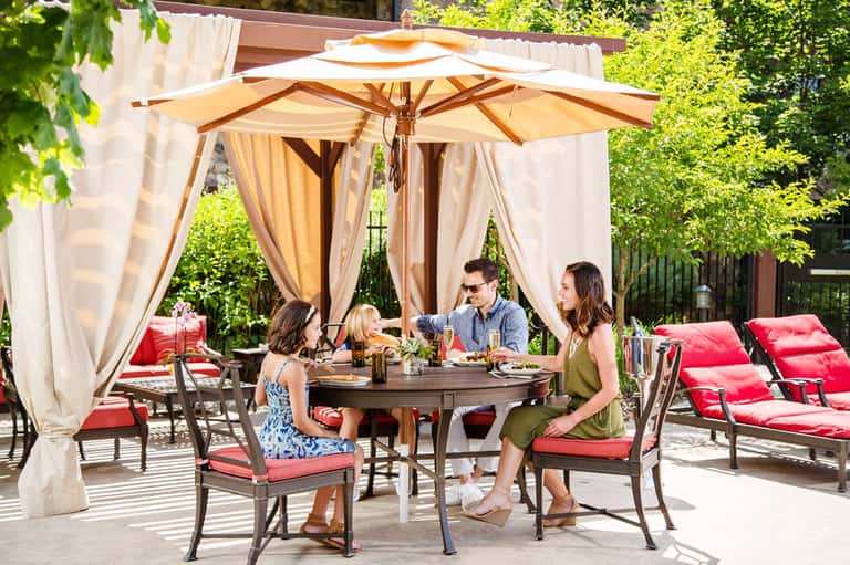 Family dining under beige umbrellas and curtains on a patio with red cushioned lounge chairs