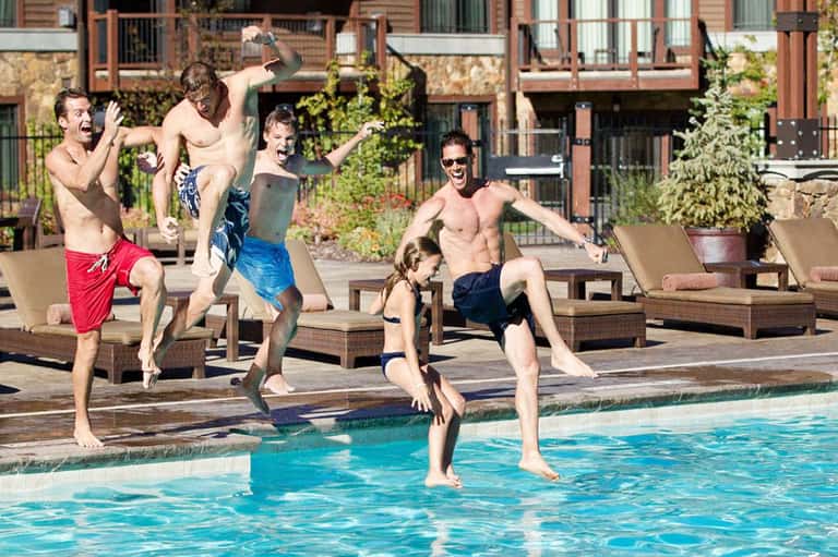 Group of men jumping into a resort pool with lounge chairs and brick buildings in background
