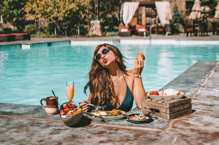 Woman in swimsuit dining poolside with fresh fruit, champagne, and tropical plants in background