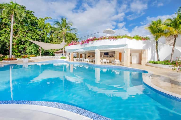 Curved resort pool with turquoise water, white lounge structure, pink flowers, and palm trees