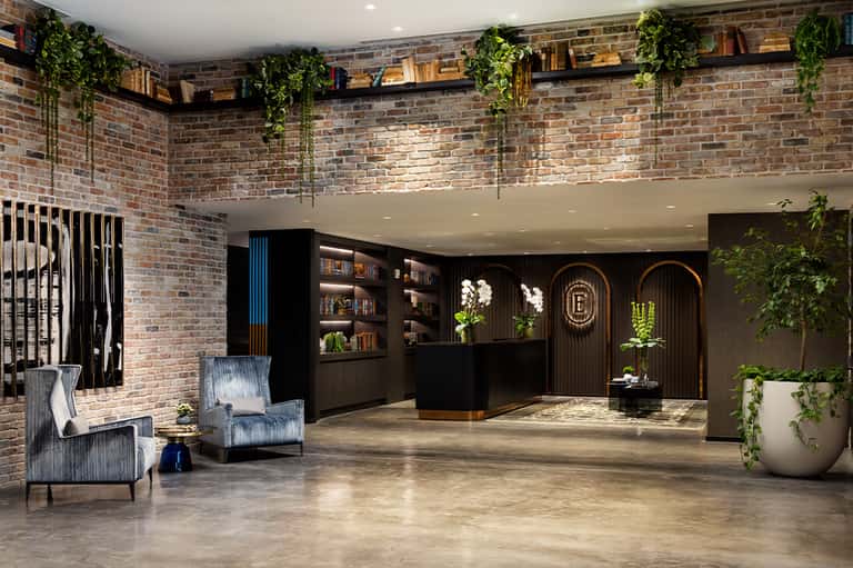 Contemporary hotel lobby with exposed brick walls, cascading plants, black reception desk, and modern furnishings