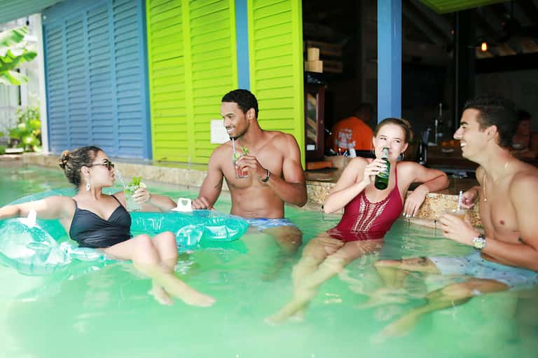 Group of four guests socializing in a turquoise pool with drinks, wearing casual resort attire in tropical setting