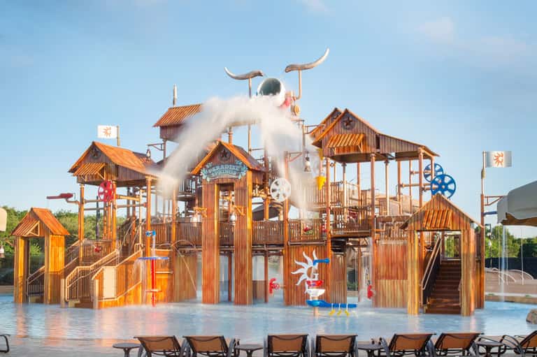 Wooden water park structure with white water slide and bull horns, surrounded by shallow pool and lounge chairs