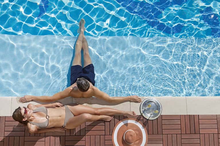 Man floating in crystal-clear pool while woman relaxes poolside with hat and refreshment