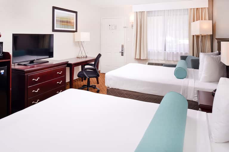 Hotel guest room with two queen beds, TV, desk, and seating area in neutral tones