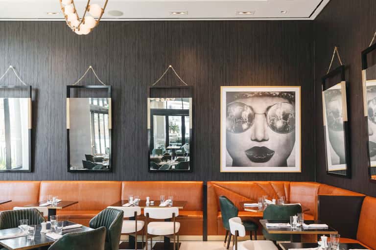 Modern restaurant interior with dark wood walls, orange booth seating, mirrors with rope accents, and contemporary art