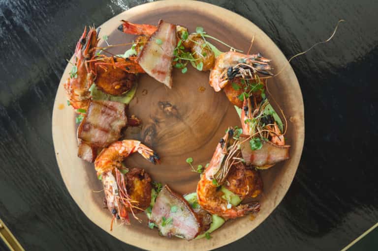 Overhead view of grilled shrimp and bacon appetizers arranged on a round wooden plate