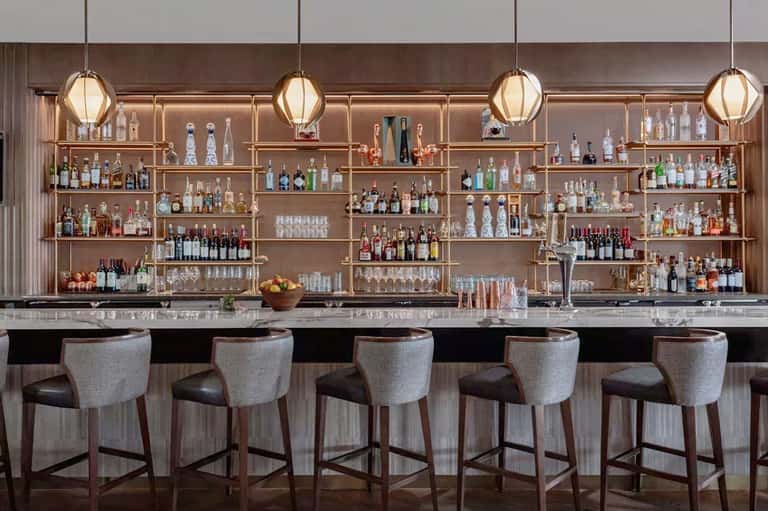 Modern bar counter with extensive liquor bottle display on wooden shelving, pendant lighting, and upholstered seating
