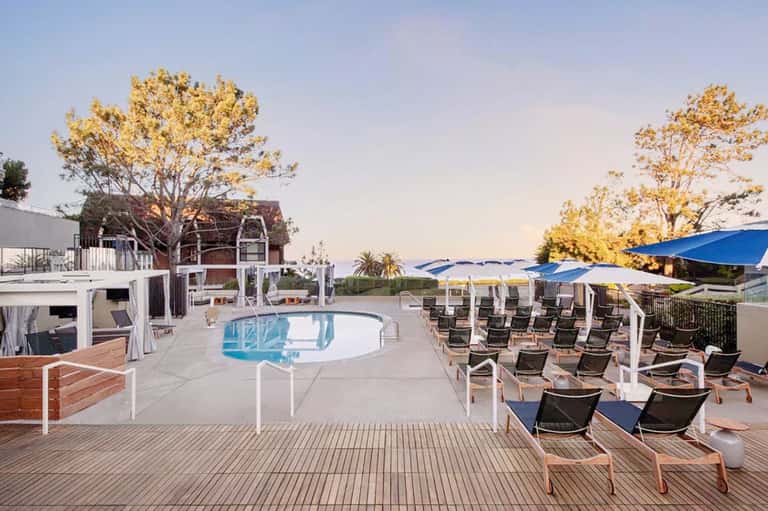 Modern pool area with white cabanas, blue umbrellas, mature trees, and lounge seating at dusk