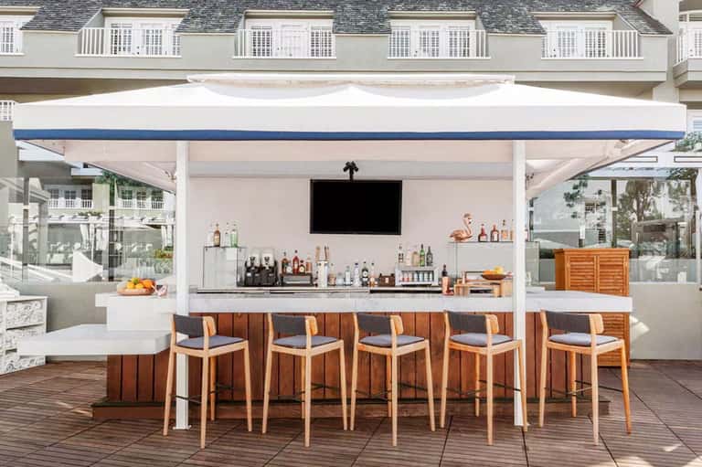 Beachfront bar with white pergola, striped awning, wooden barstools, and ocean-view seating