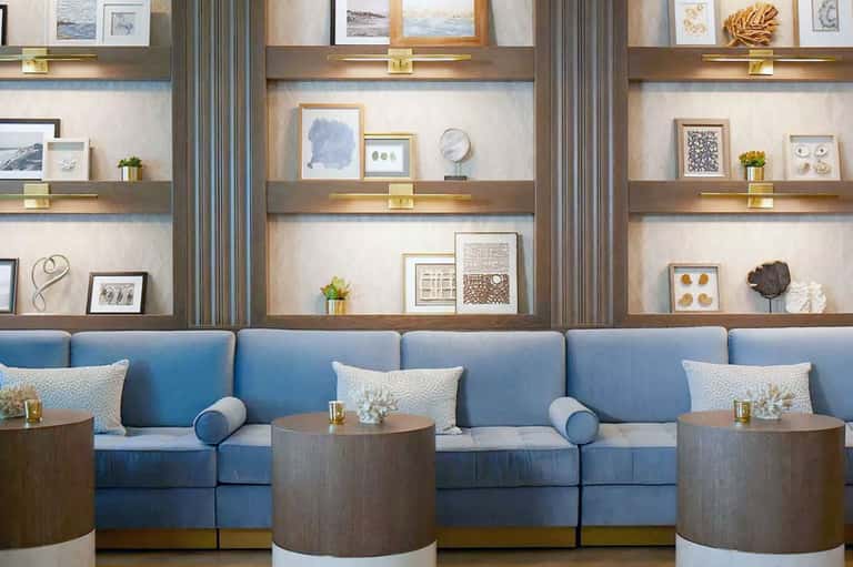 Hotel lobby lounge with blue sofa seating, wooden tables, and shelving with coastal decor