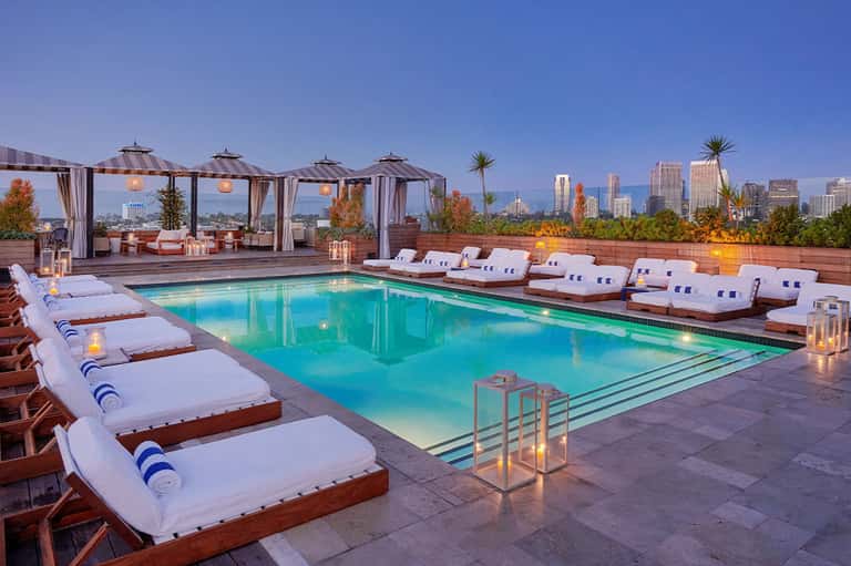 Elevated rooftop pool at dusk with lounge beds, city skyline, and ambient lighting