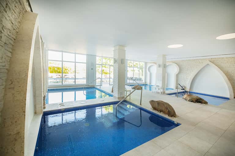 Indoor thermal spa facility with multiple blue pools, columns, and large windows overlooking exterior