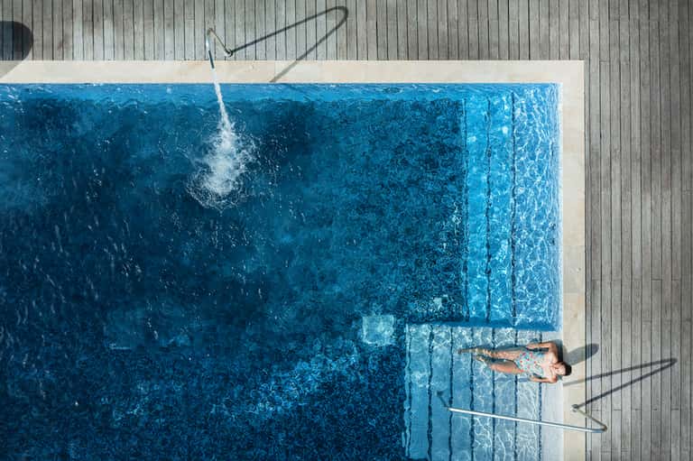 Overhead view of person swimming in bright turquoise infinity pool on wooden deck