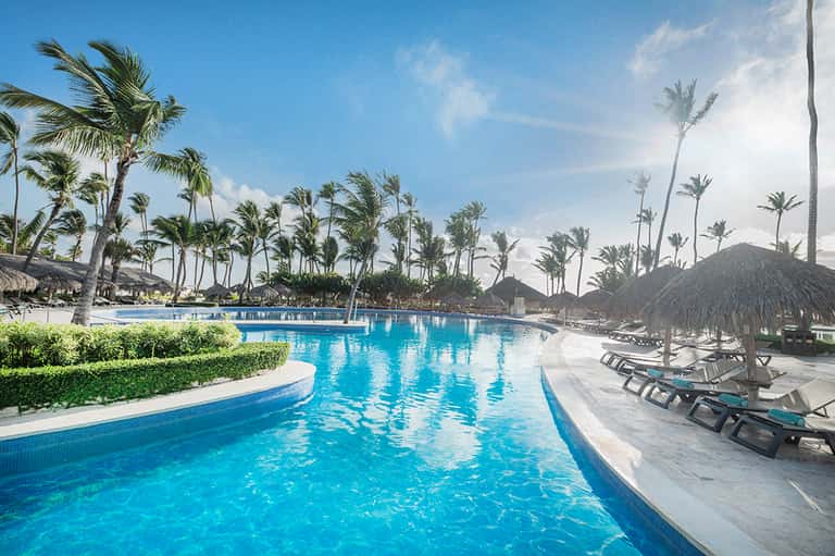 Sprawling tropical resort pool with palm trees, thatched umbrellas, palm landscaping, and ocean views