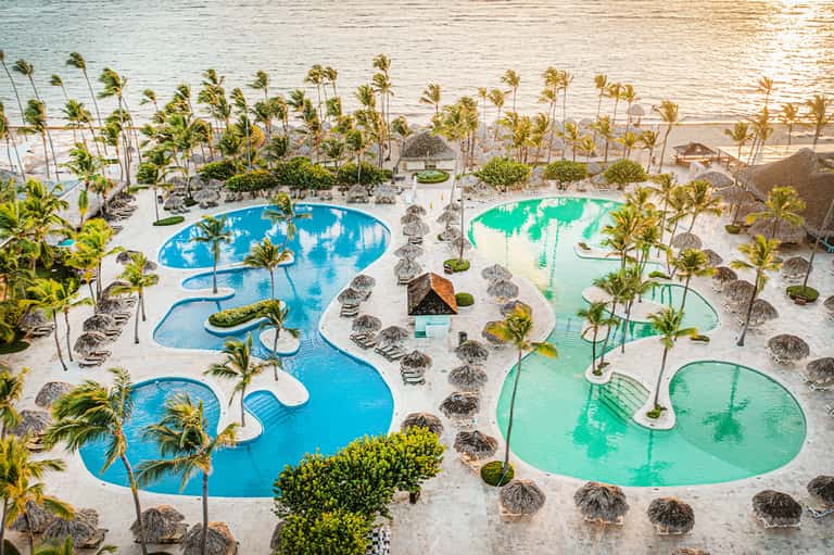 Aerial view of tropical resort pools with beach umbrellas, palm trees, and ocean waterfront