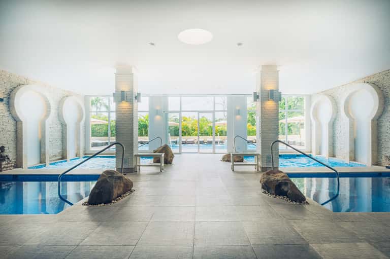 Modern spa indoor pool with symmetrical arched alcoves, decorative stone elements, and floor-to-ceiling windows